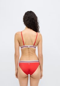 Σετ κόκκινου μπικίνι με λεπτές τιράντες, ριγωτή ταινία στο πίσω μέρος, λεία υφή και εσώρουχο με χαμηλή μέση. Λεπτομέρεια με μεταλλικό κλείσιμο.