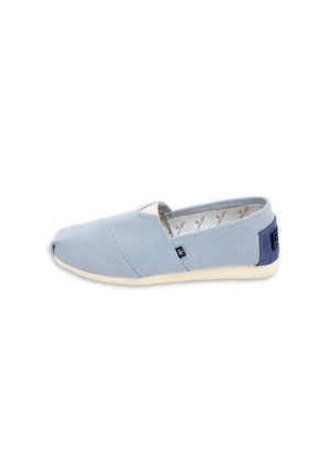 Zapato casual azul claro tipo slip-on con suela beige, parte superior de tela y parche en el talón azul marino, mostrado sobre un fondo blanco.