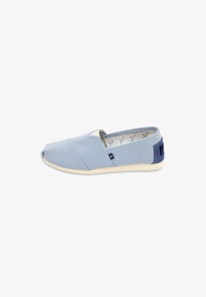 Zapato casual azul claro tipo slip-on con suela beige, parte superior de tela y parche en el talón azul marino, mostrado sobre un fondo blanco.
