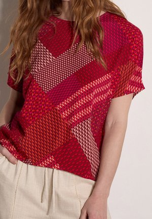 Femme portant un haut à manches courtes avec un motif patchwork géométrique rouge et rose et un pantalon beige clair à cordon.