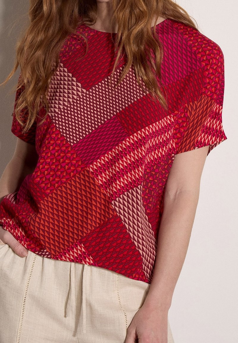 Femme portant un haut à manches courtes avec un motif patchwork géométrique rouge et rose et un pantalon beige clair à cordon.