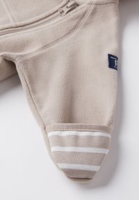 Helles beiges Fleece-Jumpsuit mit einem Reißverschluss vorne, ausgestattet mit einem gestreiften Bündchen in Beige und Weiß sowie einem kleinen Markenpatch.
