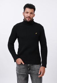 Suéter negro de punto con cuello alto y patr�ón texturizado, que presenta un pequeño logo amarillo. Combinado con jeans gris desgastados para un look informal.