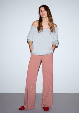 Junge Frau mit langen braunen Haaren trägt ein graues Schulterfrei-Top, rot-weiß gestreifte, weit geschnittene Hose und rote Schuhe und steht vor einer schlichten Wand.