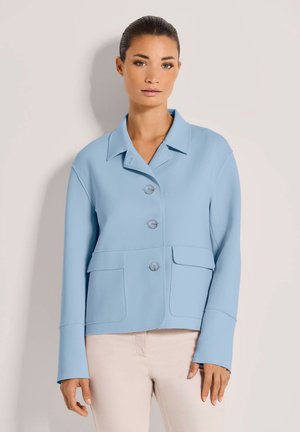 Vrouw met een lichtblauwe jas met knopen en grote zakken, en beige broek, staande tegen een eenvoudige lichte achtergrond.