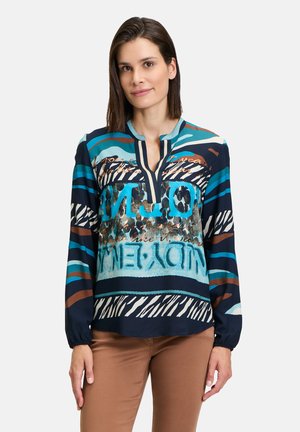 Blusa multicolore caratterizzata da una combinazione di motivi blu, marrone e bianco, con scollatura a chiave e maniche lunghe. Realizzata in tessuto morbido.
