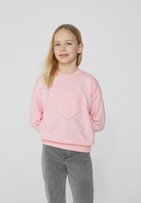 Růžová mikina s aplikací ve tvaru srdce uprostřed, vyrobená z měkkého materiálu s elastickými manžetami a lemem. V kombinaci s šedými džíny.