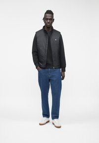 Schwarze gesteppte Weste über einem schwarzen Sweatshirt, kombiniert mit blauen Denim-Jeans und weißen Sneakers mit braunen Sohlen. Für einen lässigen Look entworfen.