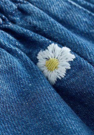 Tkanina dżinsowa w średnim niebieskim odcieniu z haftowaną białą stokrotką z żółtym środkiem, prezentująca teksturowaną powierzchnię.