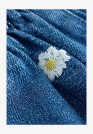 Vidējas zilas krāsas džinsa audums ar izšūtu baltu nātres ziedu dzeltenā viducī, demonstrējot teksturētu virsmu.