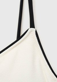 White bralette with black trim, featuring a triangular shape, smooth texture, and adjustable black straps. Stitching detail is visible.