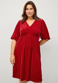 Rood fluwelen jurk met een V-hals, korte pofmouwen en een gathering taille. Zachte textuur en gladde afwerking, knie-lange silhoeet.