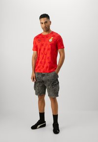 Rød grafisk t-shirt med gentagne symboler og et fodbolddesign, parret med grå cargo shorts og sorte sneakers.