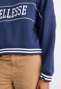 Sweat-shirt bleu marine avec passepoil blanc et lettrage, associé à un pantalon beige, montrant la partie médiane et le bras droit avec des détails sur les poignets.