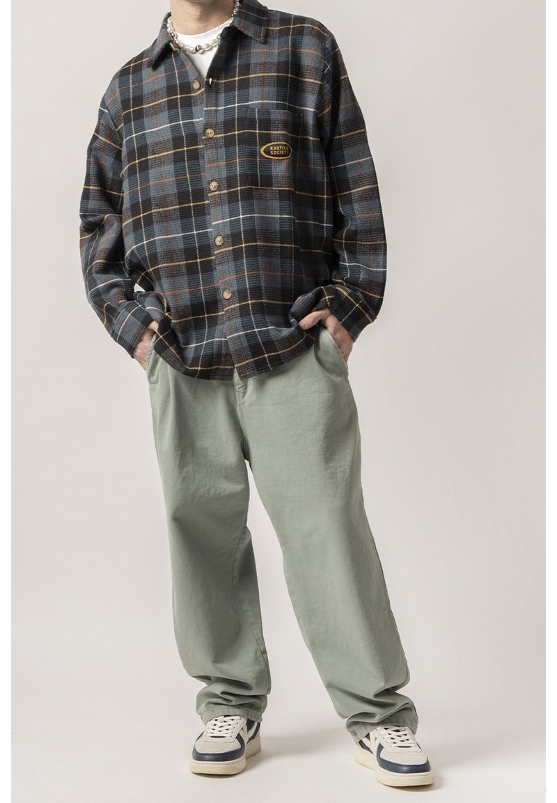 Personne portant une chemise ample à carreaux foncés boutonnée, un pantalon large vert clair et des baskets blanches avec des accents bleu marine, les mains dans les poches.