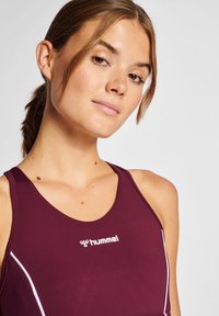 Bordowy stanik sportowy wykonany z elastycznego materiału, z białymi akcentami i logo z przodu. Zaprojektowany z dekoltem w łódkę i szerokimi ramiączkami.