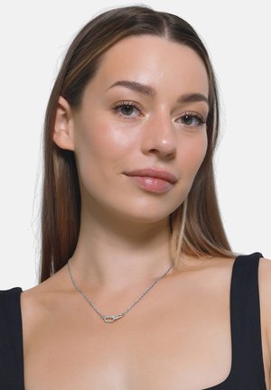 Sølvkjede med en vridd kjede og liten oval anheng, som har en jevn finish og minimalistisk design, båret på en bar hals.