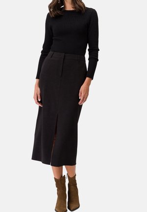 Kobieta ubrana w czarny dzianinowy top z długim rękawem, czarną midi spódnicę z przednim rozcięciem oraz brązowe zamszowe botki, stojąca na białym tle.