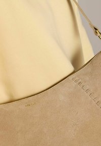 Borsa a spalla beige in suede con una texture liscia, forma curvata e dettagli cuciti. Accessori in oro con il logo "MNFLD" impresso sul lato.