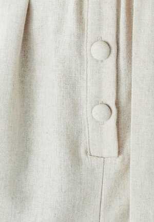 Tessuto beige chiaro con una trama texturizzata, caratterizzato da due bottoni rivestiti di tessuto sulla parte anteriore e un piccolo dettaglio di cucitura verticale.