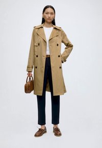 Woman wearing a beige trench coat, white top, black pants, brown belt and shoes, holding a small brown handbag, standing against white background.