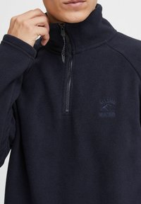 Sudadera de forro polar en color navy con cierre de media cremallera, cuello alto y logo bordado en el pecho. Textura suave y ajuste moderno.