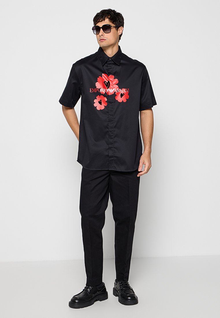Hombre con camisa negra de manga corta con flores rojas y texto, pantalones negros, zapatos negros y gafas de sol oscuras, de pie contra una pared blanca.