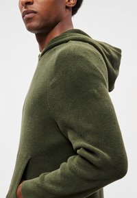 Felpa verde con cappuccio in tessuto strutturato, dotata di tasca a marsupio e spacchi laterali per facilitare i movimenti.