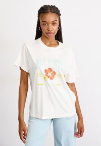 T-shirt en coton couleur crème avec un graphique coloré d'hibiscus et le texte "SUN KISSED 1990 Roxy". Manches courtes, coupe décontractée, texture douce.