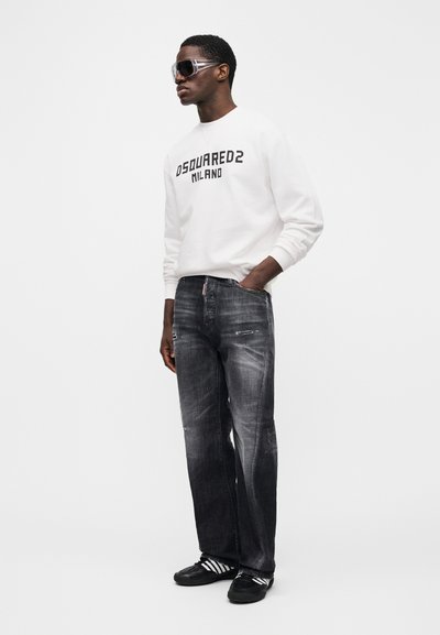 Άνδρας που φοράει λευκό φούτερ Dsquared2 Milano, ξεθωριασμένο μαύρο τζιν, μαύρα αθλητικά παπούτσια Adidas και μεγάλα διάφανα γυαλιά ηλίου, στέκεται με ένα χέρι στην τσέπη.