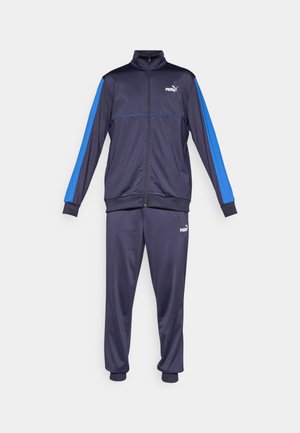 Tumši zils Puma sporta tērps ar pilna garuma jaku, kurai ir zilas svītras uz piedurknēm, un saskaņotas bikses, abiem ir Puma logotips pa kreiso sānu.