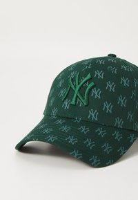 Grön keps i tyg med ett upprepat logomönster och en upphöjd logotyp framtill. Böjd skärm och justerbar rem bak.