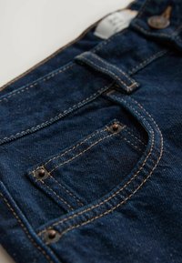 Des jeans en denim bleu foncé présentent une poche avant avec des coutures dorées contrastantes et des accessoires en bronze, mettant en avant une finition texturée.