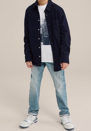Kind trägt eine marineblaue Jacke mit Knöpfen, ein weißes T-Shirt mit Grafik, hellblaue Jeans und weiße Turnschuhe und steht vor einem beigen Hintergrund.