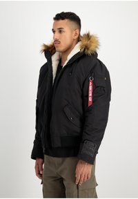 Zwarte geïsoleerde jas met nepbont capuchon, ritssluiting aan de voorkant en meerdere zakken; heeft een patch op de mouw en een rode ritslabel.
