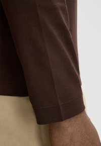 Braunes Langarmshirt mit glatter Textur, das über eine eng anliegende Manschette und subtile Nähte in einem leicht taillierten Arm verfügt.