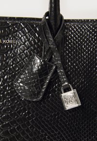 Sac fourre-tout en cuir noir avec un motif de écailles texturé. Dispose d'une étiquette amovible, d'une bandoulière tressée et d'un accent logo en métal brillant.