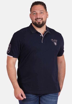 Homme souriant aux cheveux courts foncés et barbe, portant un polo bleu marine et un jean bleu, sur fond blanc.