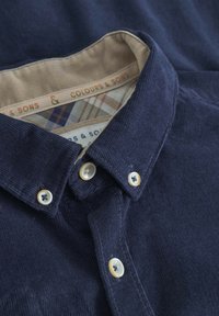 Camicia in corduroy blu navy con colletto button-down, fodera beige e cuciture decorative. Due bottoni con accenti dorati sul colletto.