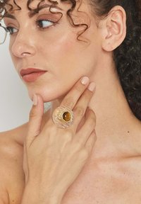 Priehľadný sklenený prsteň s drážkovanou textúrou, v strede s okrúhlym jantárovým kameňom. Ruka modelky spočíva na jej brade.