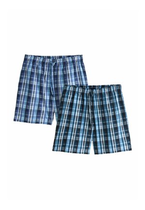 Ing nélküli fiatal férfi göndör hajjal, kék kockás rövidnadrágot viselve, mezítláb áll, kezét zsebre dugva, egy egyszerű világos fal előtt.