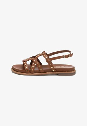 Sandalia de cuero marrón con acentos de tachuelas, múltiples correas, suela plana y correa de tobillo ajustable. Presenta una textura suave y herrajes en tono dorado.