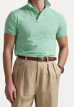 Mann trägt mintgrünes Poloshirt, das in beige plissierte Hose mit braunem Gürtel und Uhr gesteckt ist, Hand in der Tasche, neutraler Hintergrund.