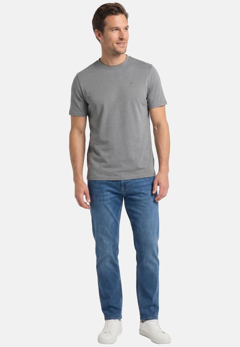 Uomo che indossa una maglietta grigia a maniche corte, jeans blu e sneakers bianche, in piedi e che guarda leggermente di lato su uno sfondo semplice.