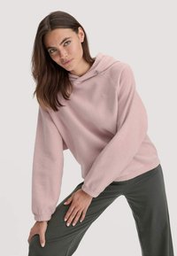 Jongeman met lang bruin haar in een lichtroze fleece hoodie en donkergrijze broek, poserend met een hand op haar knie.