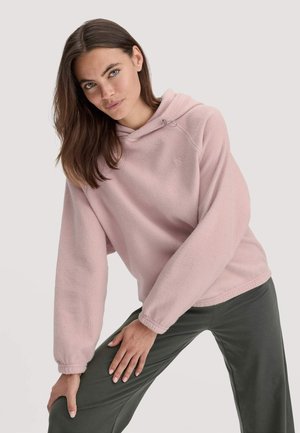 Jongeman met lang bruin haar in een lichtroze fleece hoodie en donkergrijze broek, poserend met een hand op haar knie.
