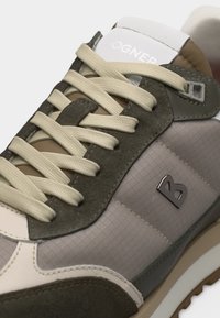 Κοντινή λήψη μπεζ και ελιάς αθλητικού παπουτσιού με πάνελ από ύφασμα και σουέντ, παχιά κρεμ κορδόνια και μεταλλικό λογότυπο "B" στην πλευρά.