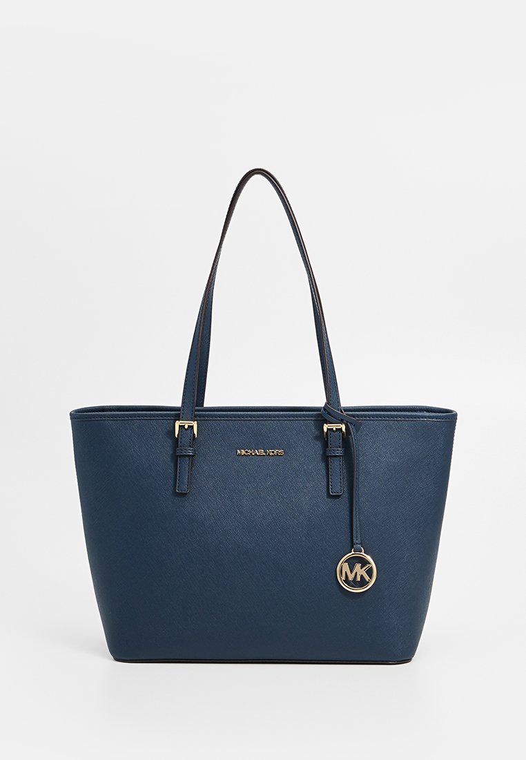 Sac fourre-tout Michael Kors bleu marine avec double sangles, plaque de logo dorée et charme rond MK sur fond blanc.