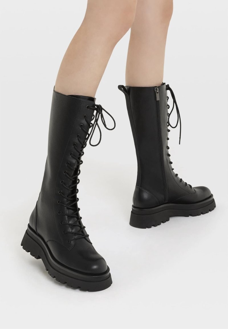 Piernas con botas negras de cuero altas, con suelas gruesas, cremalleras laterales y cordones atados en la parte superior, contra un fondo liso.