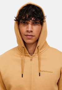 Tan zip-up hættejakke med snoretræk i hætten. Har en blød tekstur og et broderet "PeakPerformance" logo på brystet.
